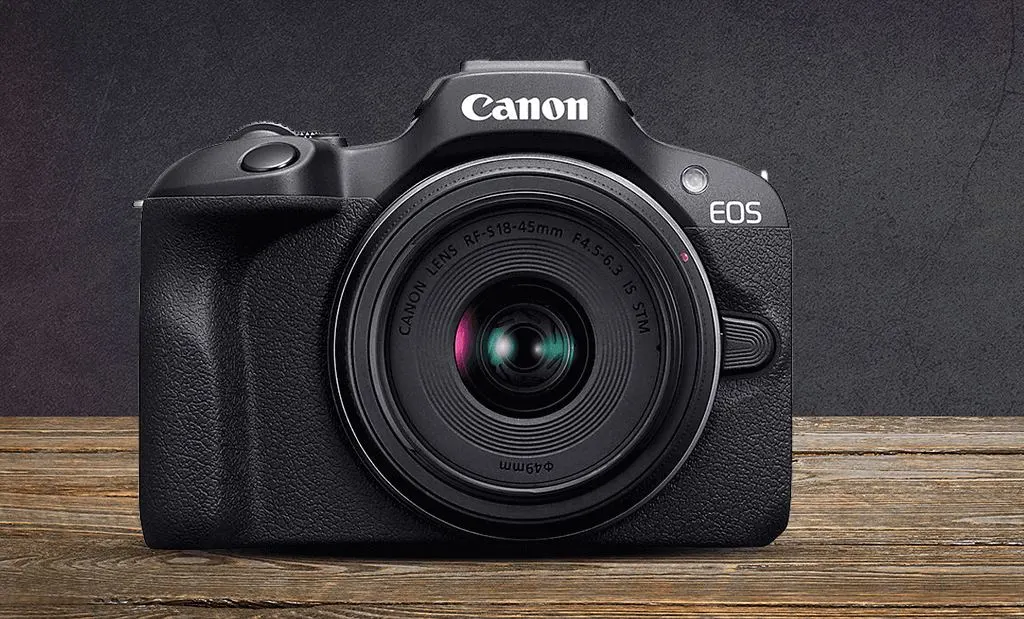 Fekete Canon EOS tükör nélküli fényképezőgép felszerelt RF-S 18-45mm objektívvel, egy rusztikus fafelületen, sötétszürke háttér előtt.