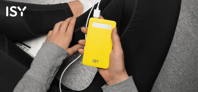 A képen egy sárga ISY power bank látható, mely egy szürke felsőt és fekete nadrágot viselő személy kezében van. Egy fehér töltőkábel csatlakozik hozzá.