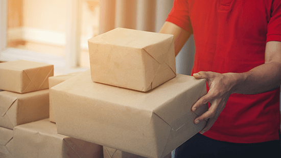 Egy piros pólós személy barna csomagokat tart, más barna csomagok halmaza mellett.