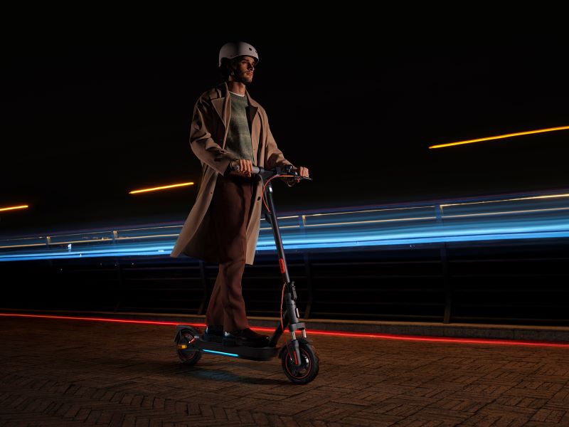 Férfi sisakban és kabátban elektromos rolleren közlekedik sötétben, fénycsíkokkal a háttérben.