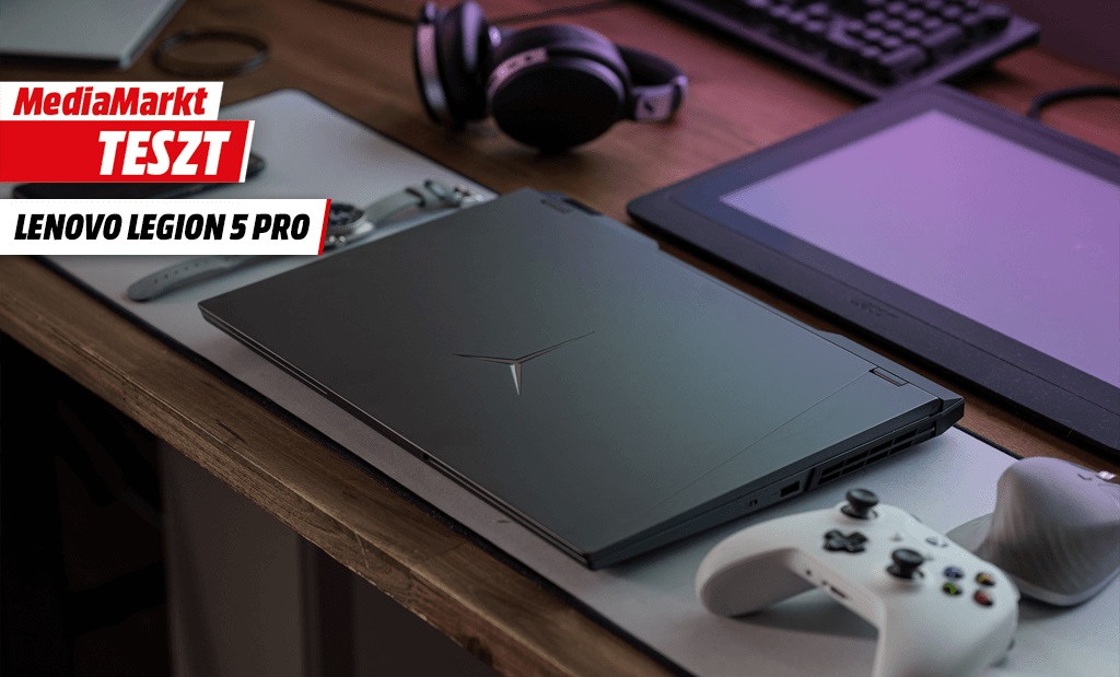 Csukott Lenovo Legion 5 Pro gamer laptop egy íróasztalon, mellette egy fehér kontrollerrel és MediaMarkt Teszt feliratú logóval.
