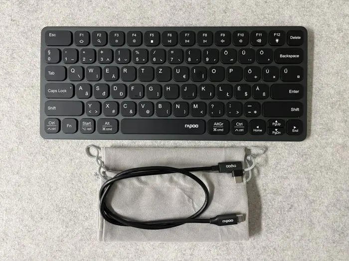Fekete, vezeték nélküli Rapoo billentyűzet szürke tartózsákkal és egy fekete USB-kábellel.