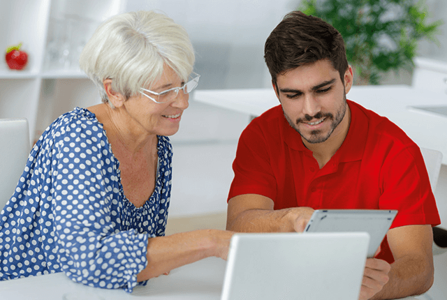 Nő kék pöttyös blúzban, férfi piros pólóban, tabletet néznek fehér asztalnál.