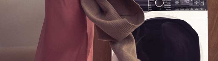 Egy piros ruhás ember tart egy barna pulóvert egy fehér mosógép előtt.