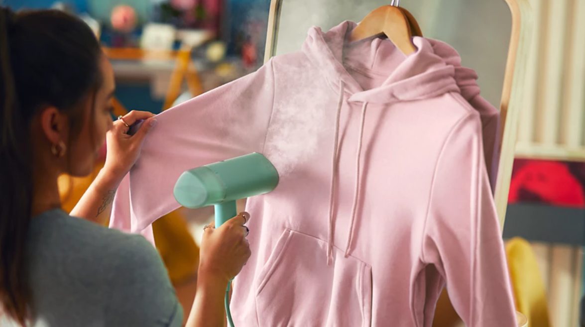 Fiatal nő rózsaszín kapucnis pulóvert gőzöl Philips 3000-es sorozatú kézi gőzölővel