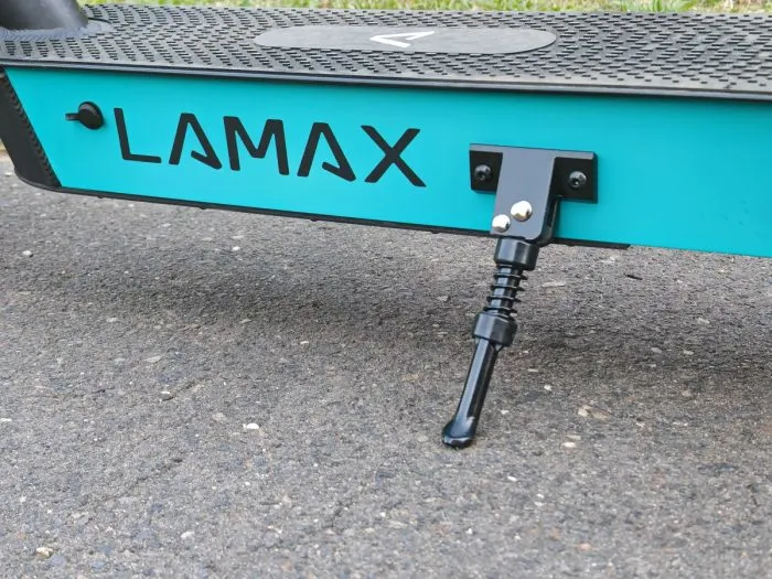 Közeli felvétel egy türkizkék színű Lamax elektromos roller vázáról és a kihajtott fekete kitámasztójáról az aszfalton.