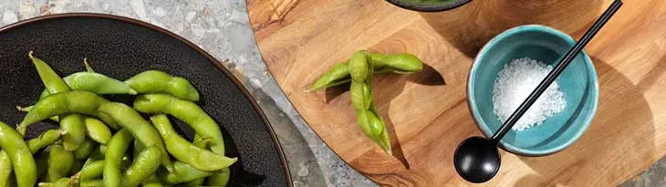 Edamame bab egy tálban és vágódeszkán. Egy kék tálban só, mellette fekete kanál.