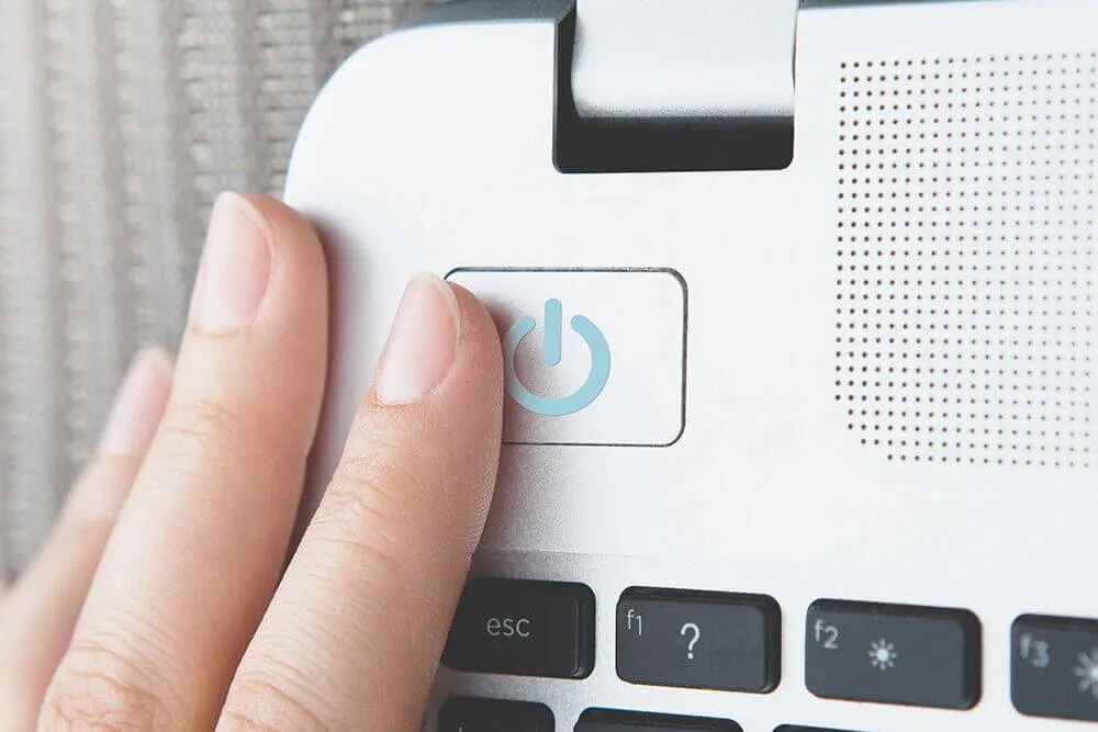 Egy kéz nyomja meg a fehér laptop bekapcsológombját, melynek közelében fekete billentyűk találhatók.