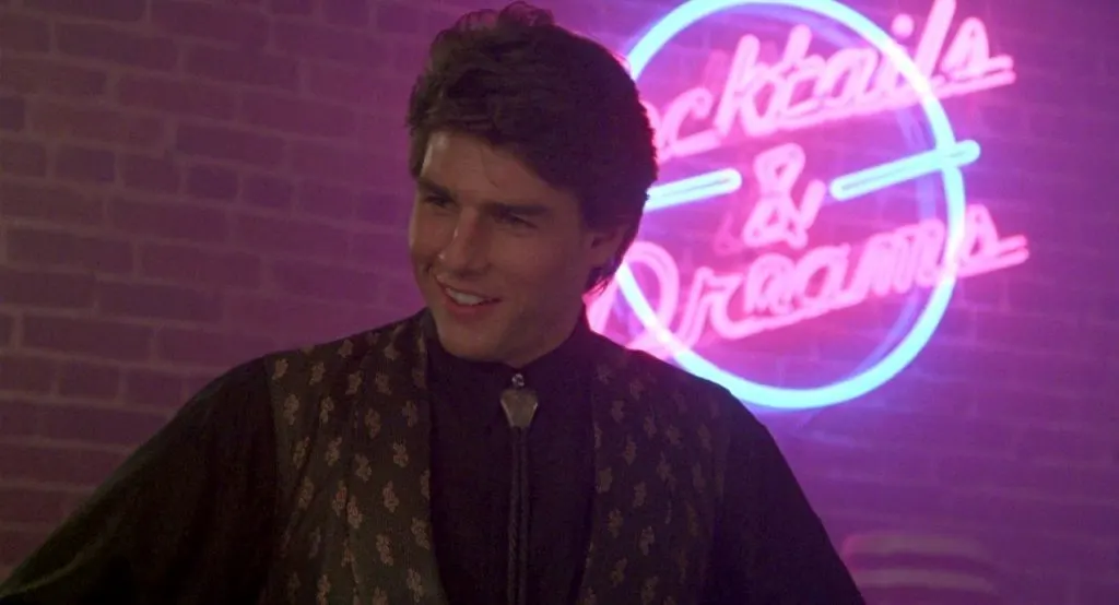 Tom Cruise mosolyog, fekete ingben és mintás mellényben, a háttérben "Cocktails & Dreams" feliratú neonfény világít.