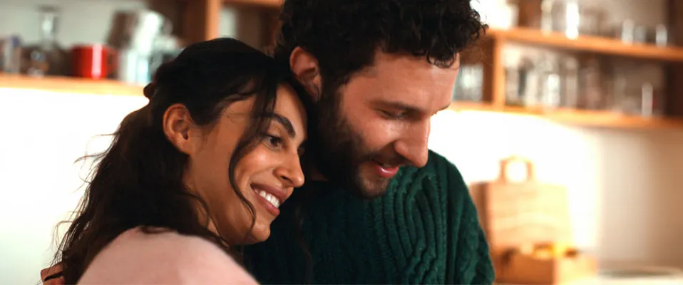 Egy mosolygó pár áll a konyhában: egy nő rózsaszín felsőben, egy férfi zöld kötött pulóverben.