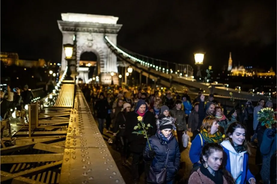 Emberek esti, virágos felvonulása a kivilágított budapesti Lánchídon, a háttérben a budai vár és a város fényei látszanak.