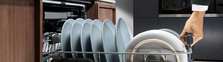 Egy kéz egy mosogatógép kosarát húzza ki, melyben kék tányérok és fehér lapostányérok vannak.