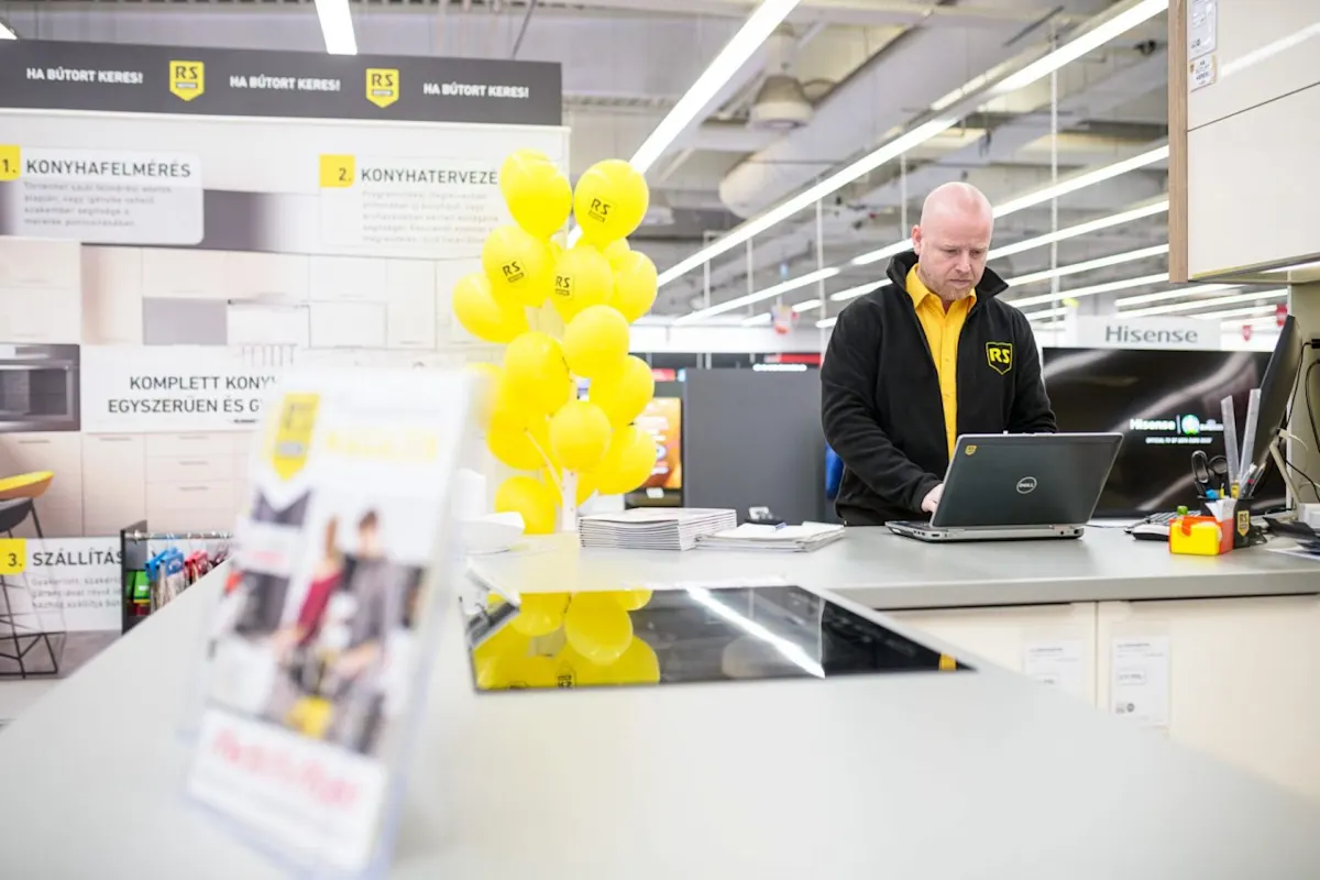 Kopasz, fekete kabátos eladó laptopon dolgozik egy bútorüzlet információs pultjánál, mellette sárga lufik és tájékoztató táblák láthatóak.