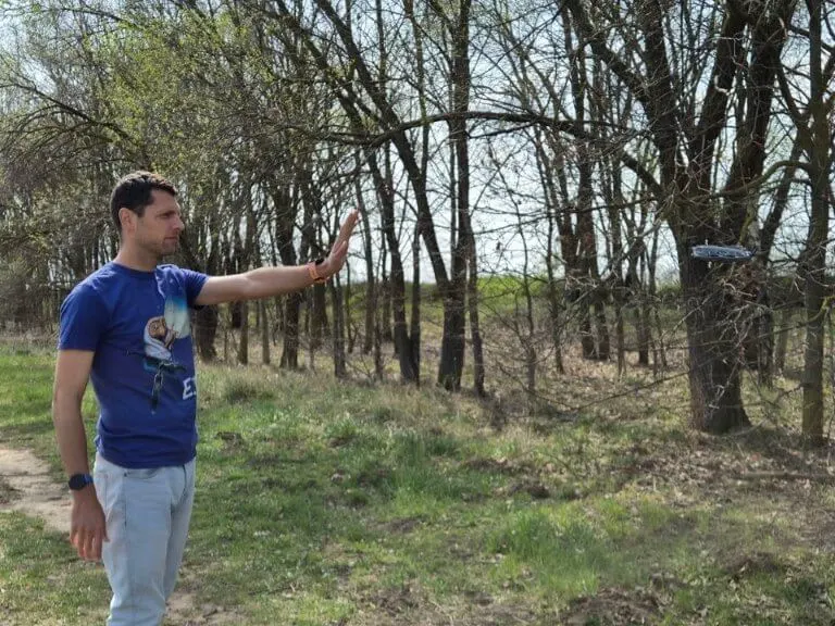Egy férfi kék pólóban egy mezőn áll és integet az előtte repülő drónnak.