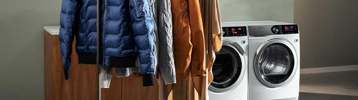 Két fehér mosógép látható a háttérben, előtérben ruhák lógnak egy állványon, barna fa szekrény mellett.