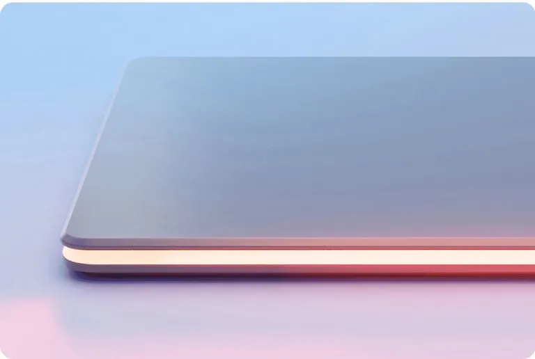 Szürke laptop sarkának közeli képe, narancssárga csíkkal a szélén, világoskék és rózsaszín háttérrel.