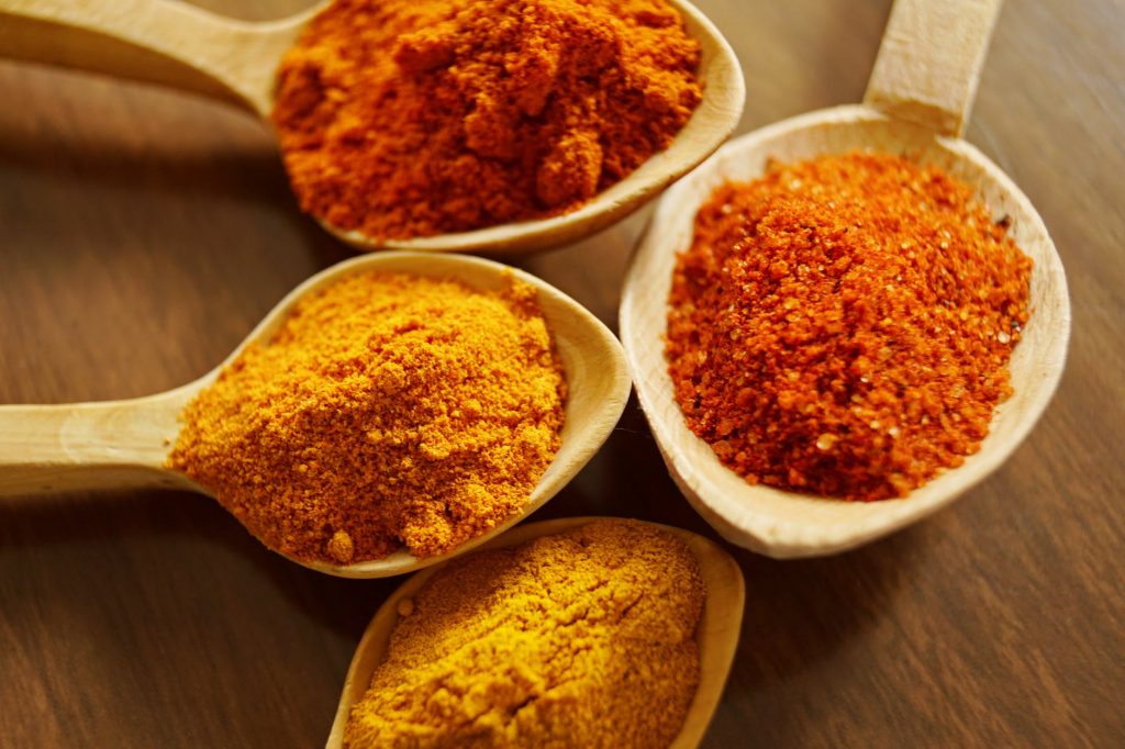 Különböző színű őrölt fűszerek, mint a paprika és a kurkuma, fa kanalakon elrendezve.