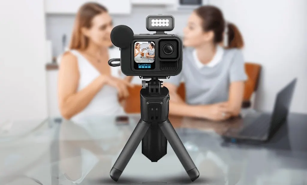 GoPro HERO13 Black Creator Edition csomag: az akciókamera állványra rögzítve, kiegészítő lámpával és külső mikrofonnal felszerelve, a háttérben két homályos női alakkal egy asztalnál.