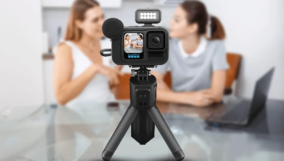 GoPro HERO13 Black Creator Edition csomag: az akciókamera állványra rögzítve, kiegészítő lámpával és külső mikrofonnal felszerelve, a háttérben két homályos női alakkal egy asztalnál.