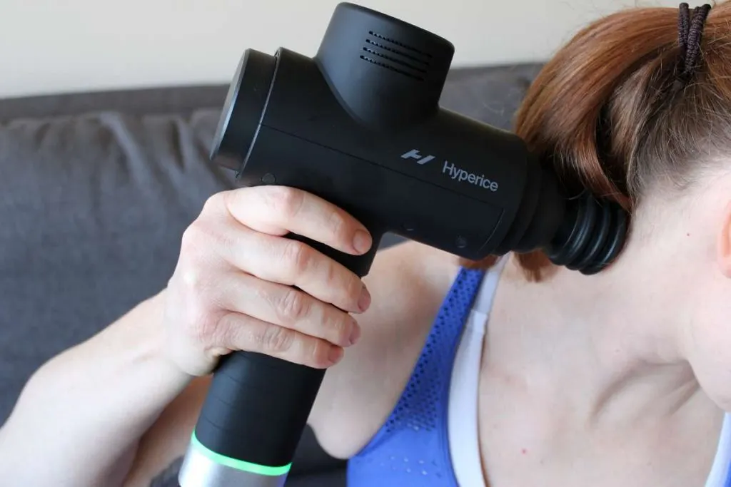 Kék trikós nő a tarkóját és a nyakát kezeli egy fekete Hyperice Hypervolt 2 Pro masszázspisztollyal.