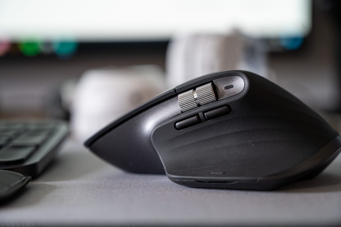 Ergonomikus prémium vezeték nélküli számítógépes egér oldalnézeti makrófotója, modern matt fekete felülettel, fém görgővel és programozható oldalgombokkal. Letisztult minimalista irodai környezet, elmosott billentyűzet és monitor a háttérben. 