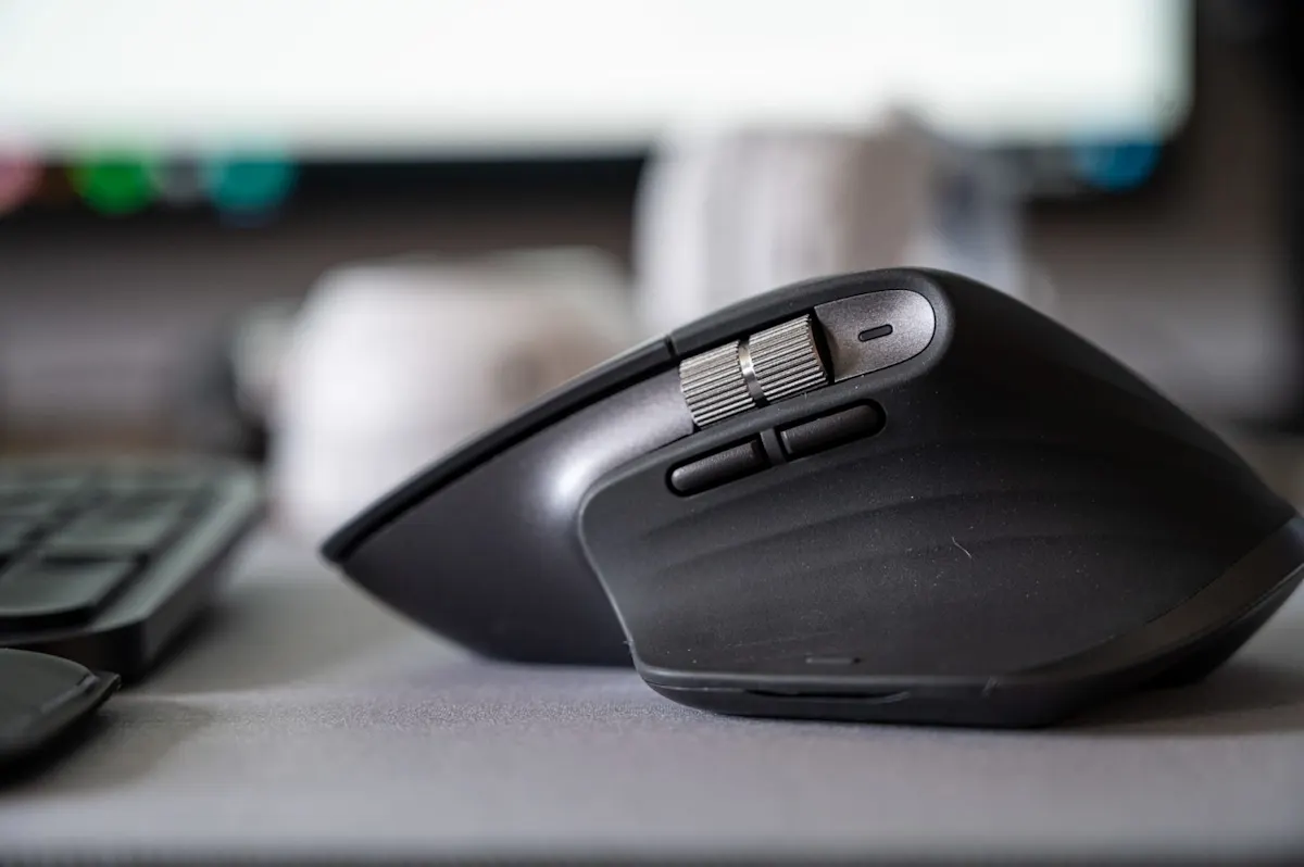 Ergonomikus prémium vezeték nélküli számítógépes egér oldalnézeti makrófotója, modern matt fekete felülettel, fém görgővel és programozható oldalgombokkal. Letisztult minimalista irodai környezet, elmosott billentyűzet és monitor a háttérben.