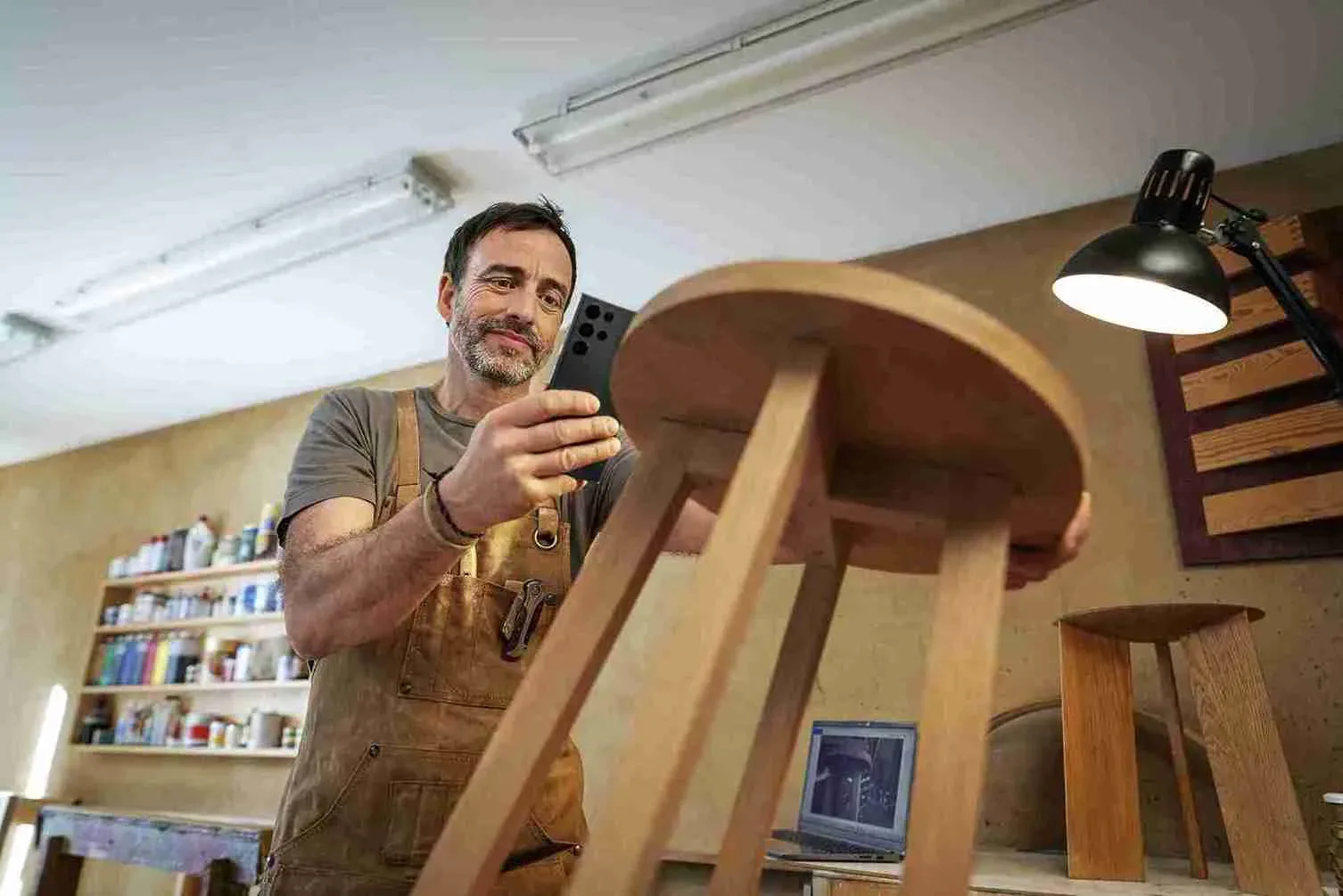Férfi műhelyben, fa kerek asztalt fotóz fekete telefonnal. Munkapadon laptop és készletek, asztali lámpa világít.