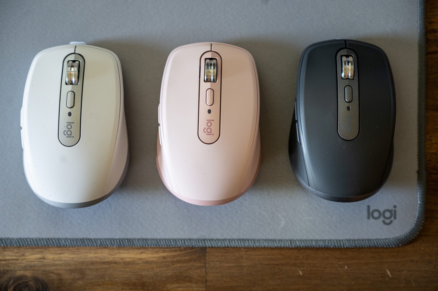 Felülnézetes termékfotó három Logitech vezeték nélküli egérről különböző színekben (fehér, rózsaszín és grafitszürke), szürke egérpadon elhelyezve fa asztalfelületen.