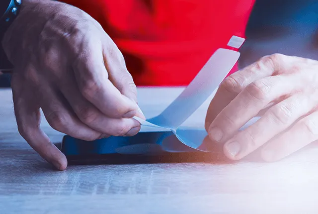 Két kéz kék telefonra helyez fel egy átlátszó fóliát, vörös háttér előtt, fa asztalon.