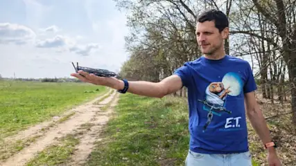 Egy férfi a tenyerében tart egy szürke drónt, miközben egy füves területen áll.