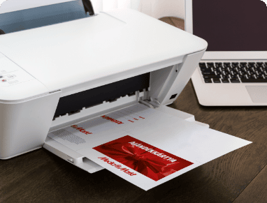 Nyomtató fehér papírt nyomtat, piros szöveggel. Laptop látható a háttérben, fekete billentyűzettel, fa asztalon.