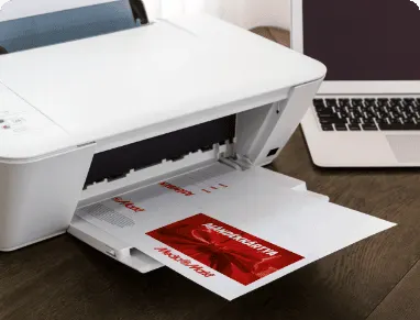 Nyomtató fehér papírt nyomtat, piros szöveggel. Laptop látható a háttérben, fekete billentyűzettel, fa asztalon.