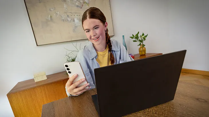 Egy mosolygó nő egy asztalnál ül laptop előtt, kezében telefont tart, a háttérben egy kép és növények láthatók.