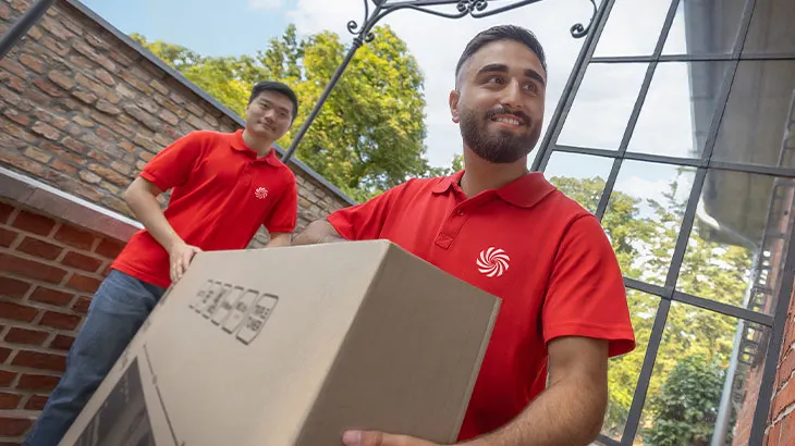 Két férfi piros pólóban kartondobozt cipel. A háttérben téglafal és zöld növényzet látható, az előtérben ablakkeret.