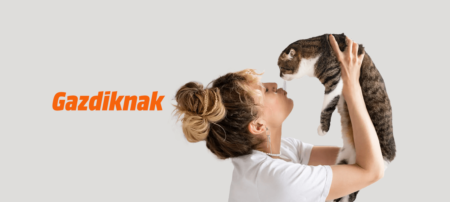 Nő tart egy csíkos macskát a szürke háttér előtt. Narancssárga "Gazdiknak" felirat látható.