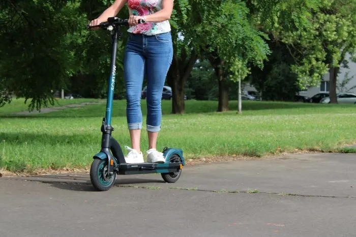 A képen látható nő egy SoFlow SO4 Pro (vagy hasonló típusú) elektromos rolleren áll egy park melletti aszfaltozott úton.