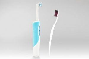 Egy fehér-kék elektromos fogkefe és egy sötét sörtéjű hagyományos fehér fogkefe egymás mellett, semleges szürke háttér előtt.