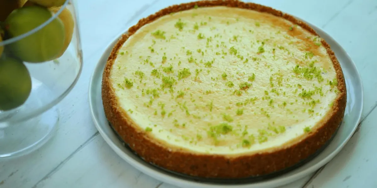 Friss, reszelt lime-héjjal díszített sült sajttorta tálcán, mellette üvegedényben citrusfélékkel.