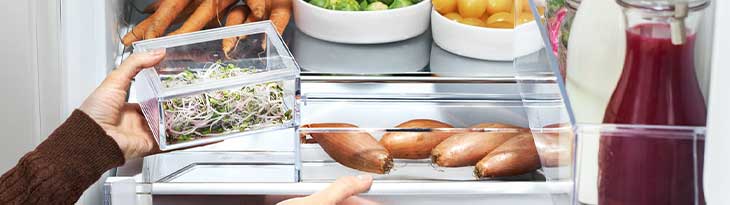 Egy hűtőszekrény belseje zöldségekkel, gyökérzöldségekkel, burgonyával és egy üveg piros folyadékkal. Egy kéz egy áttetsző edényt tart.