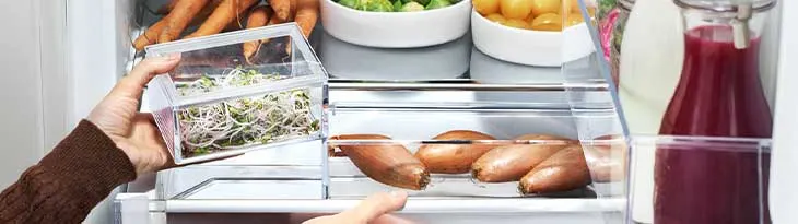 Egy hűtőszekrény belseje zöldségekkel, gyökérzöldségekkel, burgonyával és egy üveg piros folyadékkal. Egy kéz egy áttetsző edényt tart.