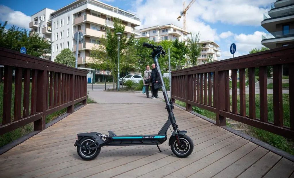 Lamax eRacer SC50 nagyteljesítményű elektromos roller oldalnézetből egy fából készült gyalogoshídon, háttérben modern városi társasházakkal, felhős kék égbolttal és járókelőkkel.