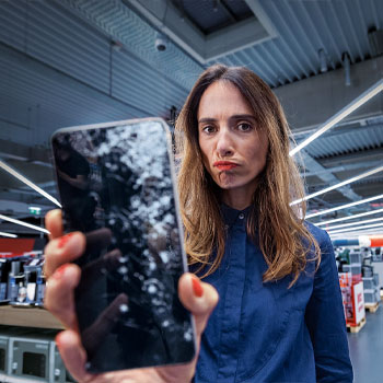 Nő kék ingben, egy törött képernyőjű okostelefont tart a kezében, egy üzlethelyiségben áll.