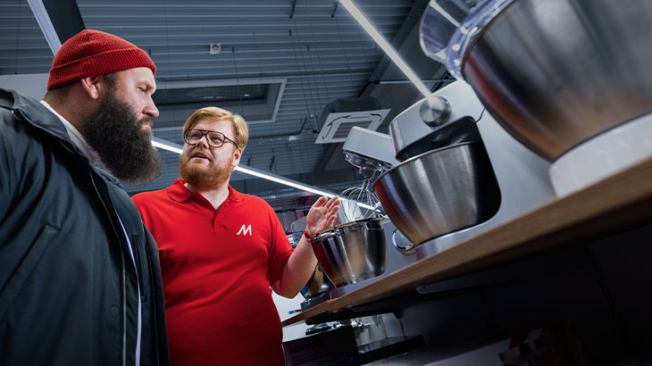 Egy vörös sapkás férfi néz egy piros pólós eladóra, aki konyhai robotgépeket mutat egy üzletben.