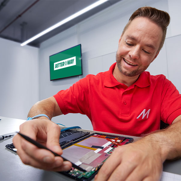 Férfi piros pólóban, "M" betűvel, tabletet javít, fekete eszközzel. A háttérben zöld képernyő, "BETTERWAY" felirattal.