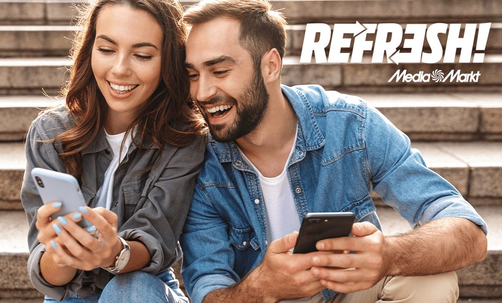 Mosolygó fiatal pár okostelefonokat néz a szabadban egy lépcsőn ülve, MediaMarkt Refresh logóval a háttérben.