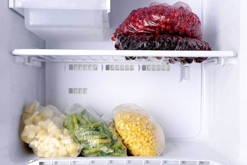 Fagyasztott élelmiszerek egy fehér fagyasztóban: piros bogyók, kukorica, zöldbab, karfiol átlátszó zacskókban.