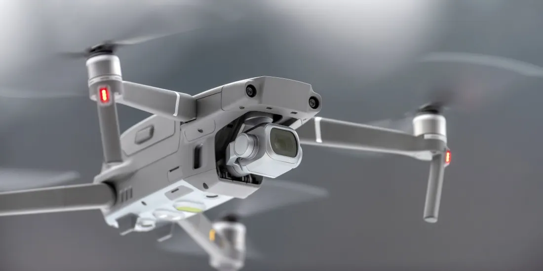 Szürke színű DJI Mavic drón repülés közben, fókuszban a beépített kamerájával és a stabilizáló gimbal egységgel.