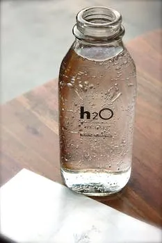 H2O feliratú, cseppkő mintázatú üvegkulacs friss, buborékos ivóvízzel egy faasztalon.