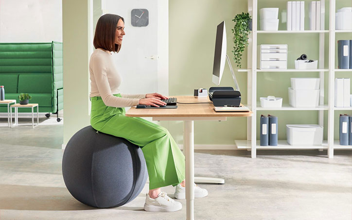 Nő ergonomikus ülőlabdán ülve dolgozik modern irodai környezetben számítógép előtt.