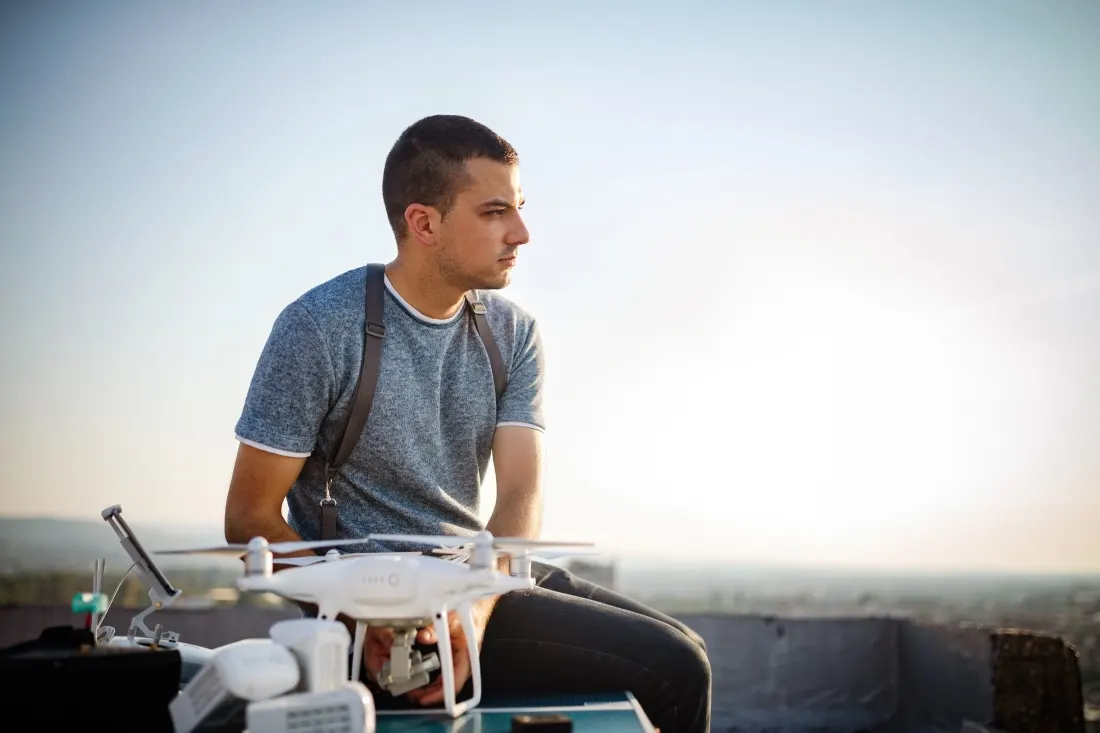 Fiatal férfi ül egy tetőn egy fehér DJI Phantom drón és a tartozékai mellett, miközben oldalra tekint a naplementében.